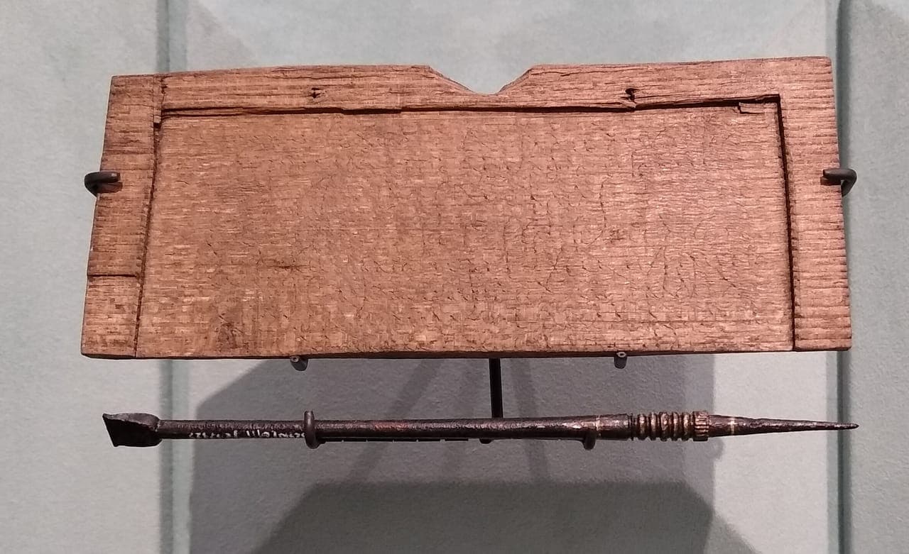 The surviving strip of the tablet on a display mount: brown wood with a V-shaped notch in its top edge and faint scratched lines, with an iron stylus shown below it