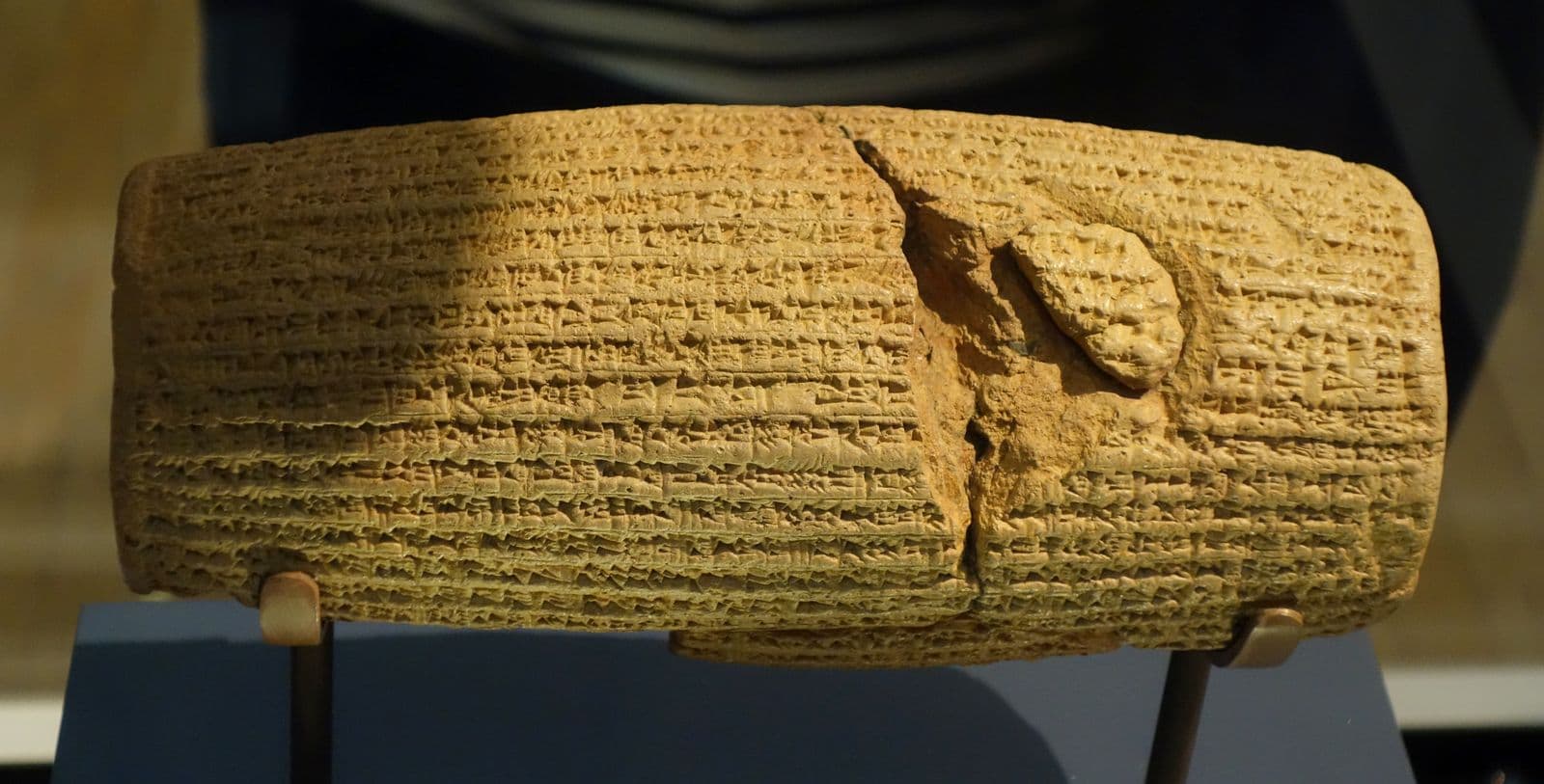 A barrel-shaped clay cylinder lying horizontally on a museum mount, its surface covered in close horizontal lines of cuneiform, with a large piece broken away from the upper right.