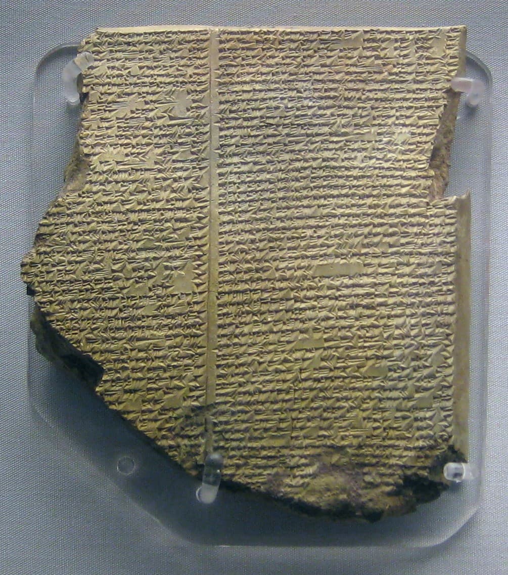 A broken clay tablet held upright by clear clips on a grey museum mount, its face packed with small cuneiform wedges in two columns divided by a vertical line. The left edge and the bottom are broken away.