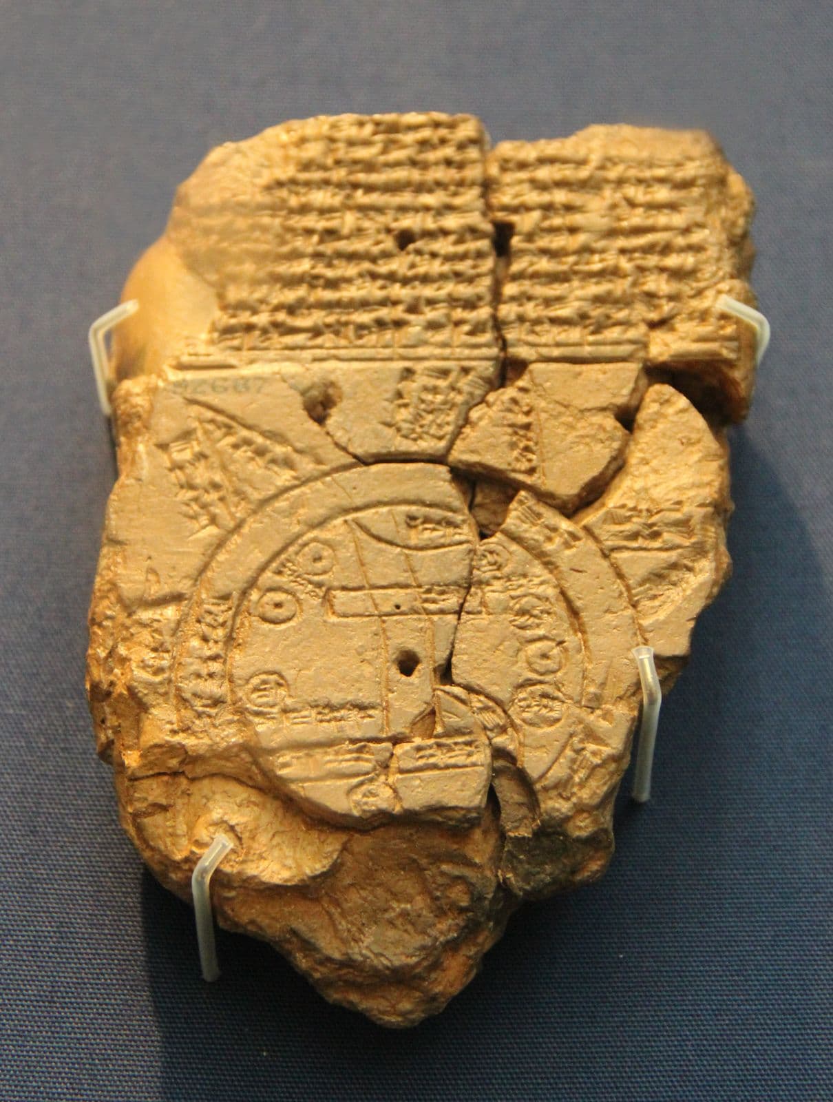A broken clay tablet held by clear clips against a blue-grey mount, the museum number 92687 painted on it: a block of cuneiform across the top, below it a double circle enclosing small rectangles and ovals, with triangles jutting out from the ring. The lower part is crumbled away.
