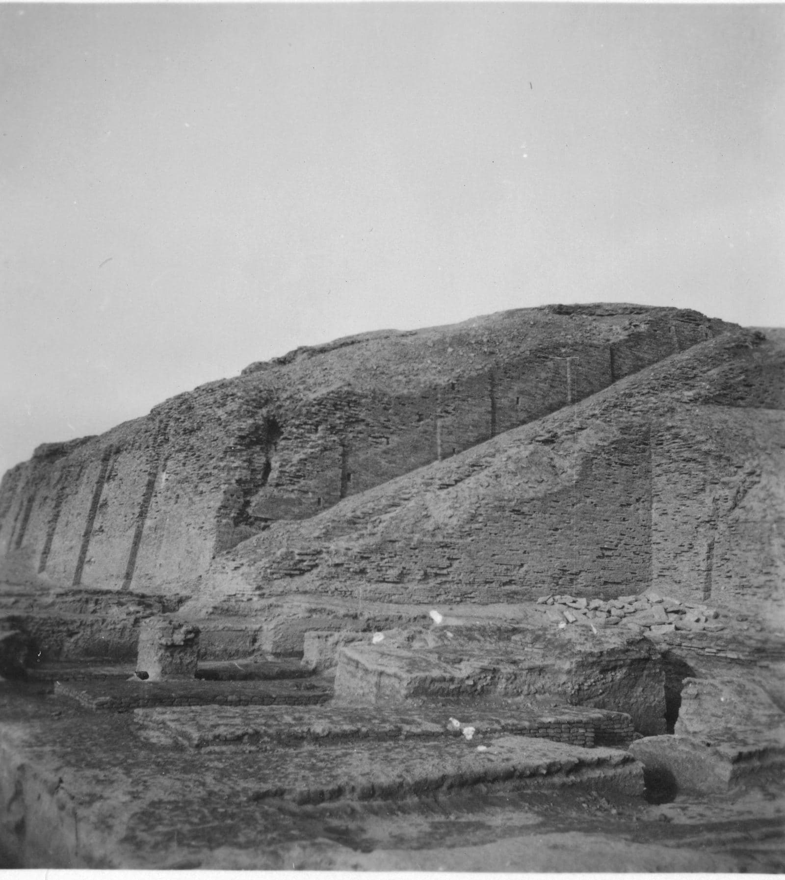 Black-and-white photograph of a large eroded mud-brick ziggurat with buttressed sides rising above excavation trenches in a flat desert