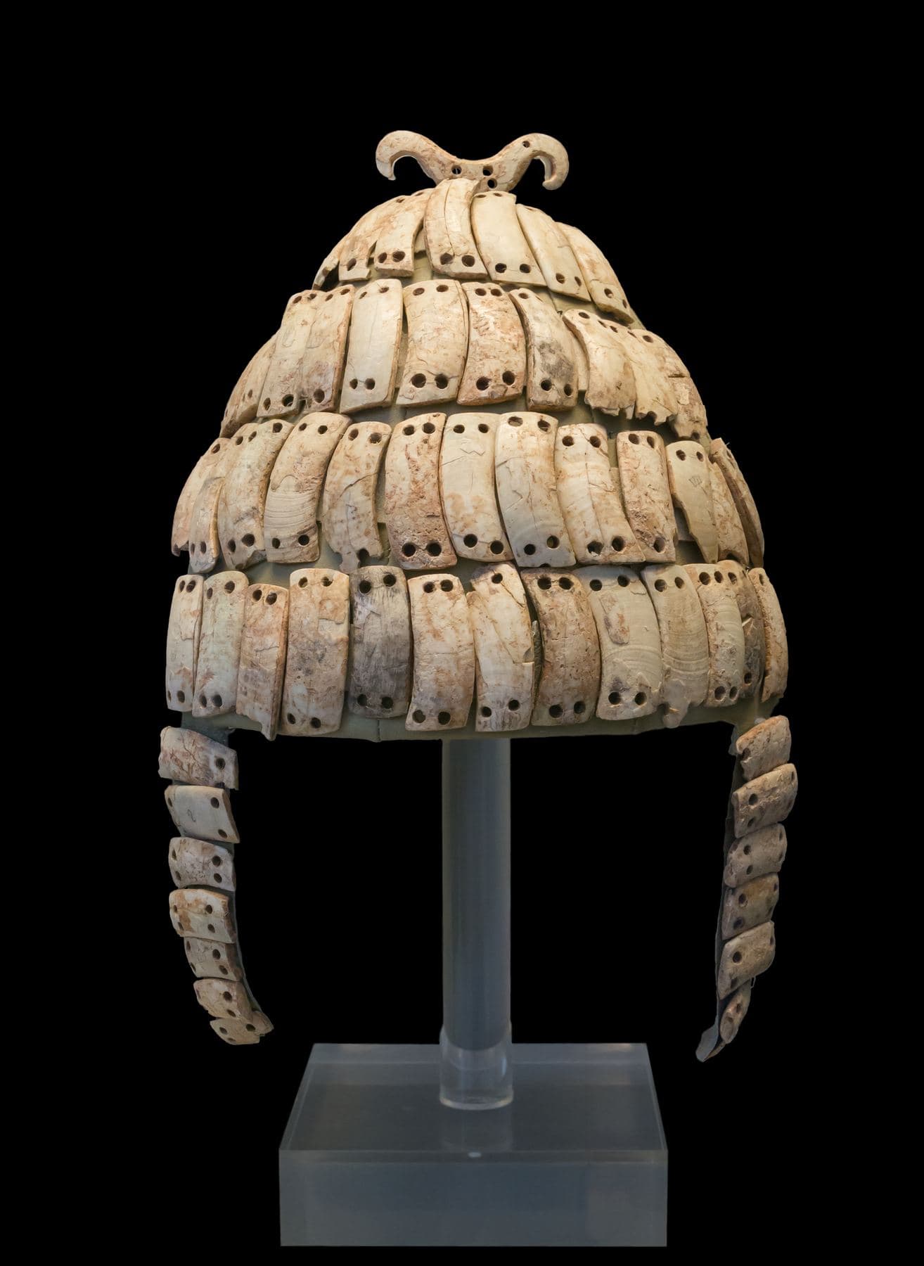 Conical helmet made of rows of curved white boar tusks stitched to a cap, with cheek-guards, on a museum stand