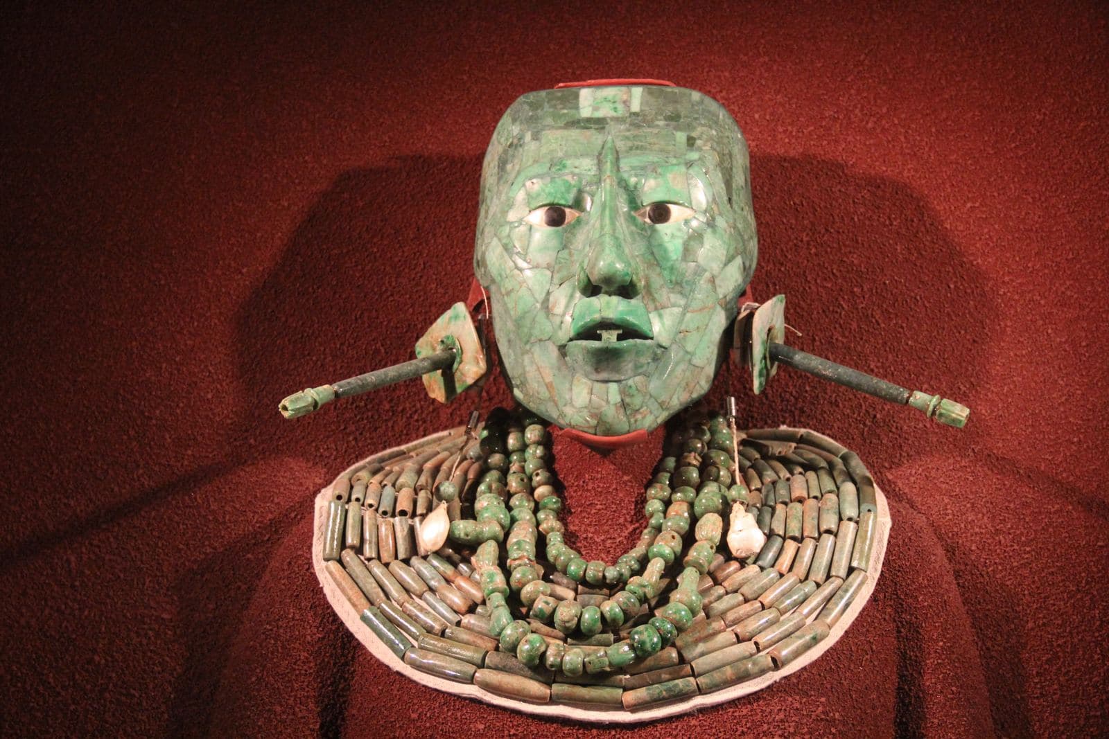 Close-up photograph of a green jade mosaic funerary mask with pale shell and dark obsidian eyes, displayed in a museum case