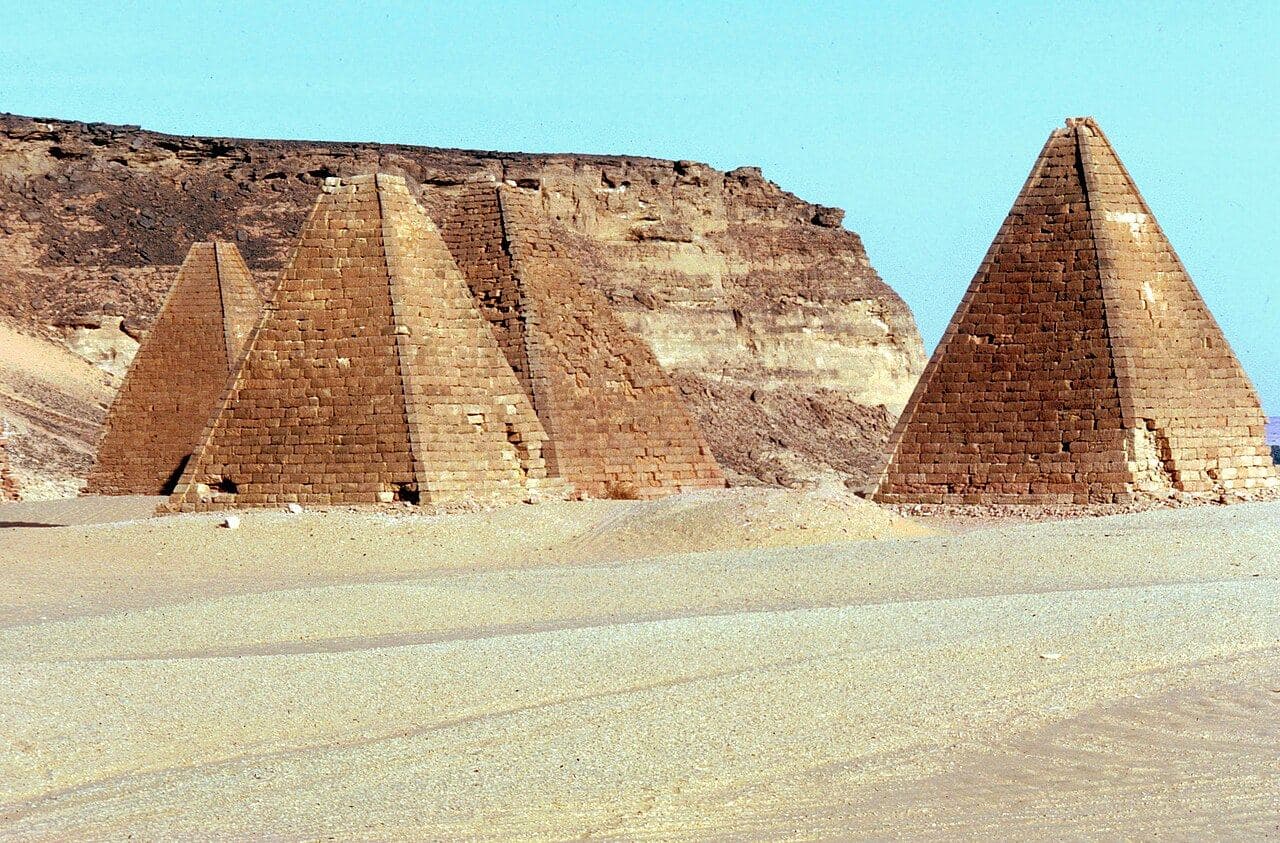 Photograph of steep-sided desert pyramids at the base of a flat-topped sandstone mountain, under a clear blue sky