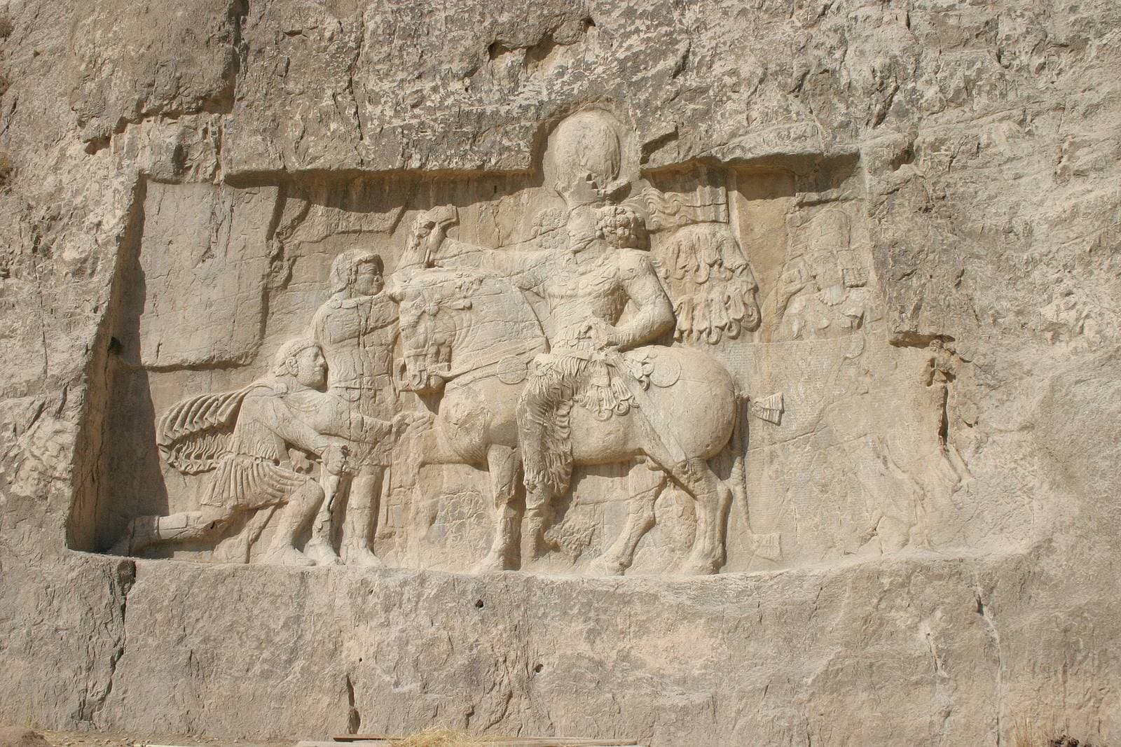 Rock-cut relief of a crowned king on horseback gripping the wrist of a kneeling captive, carved into a cliff face