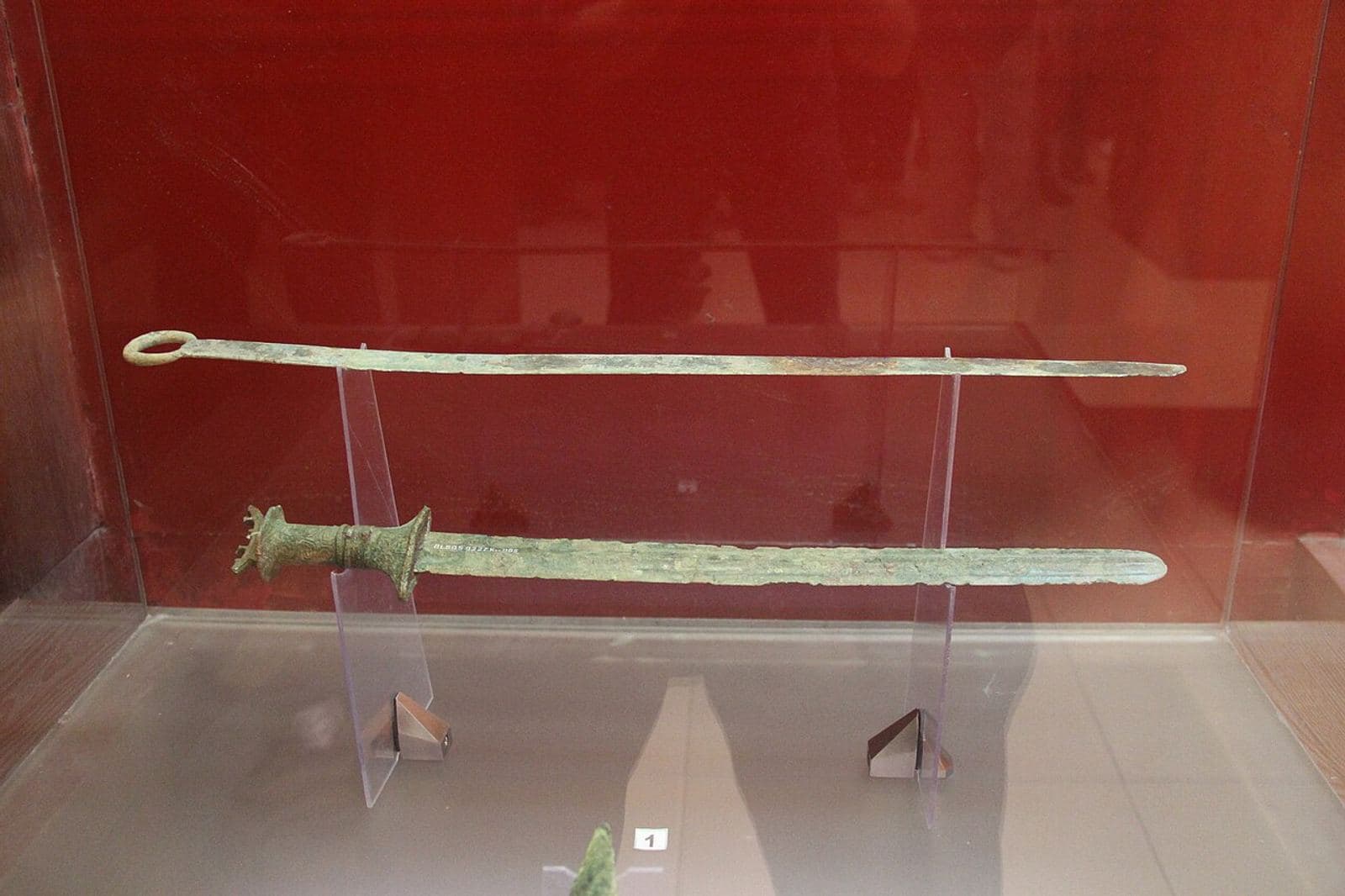 A row of bronze swords and spearheads with corroded green patina, displayed in a museum case