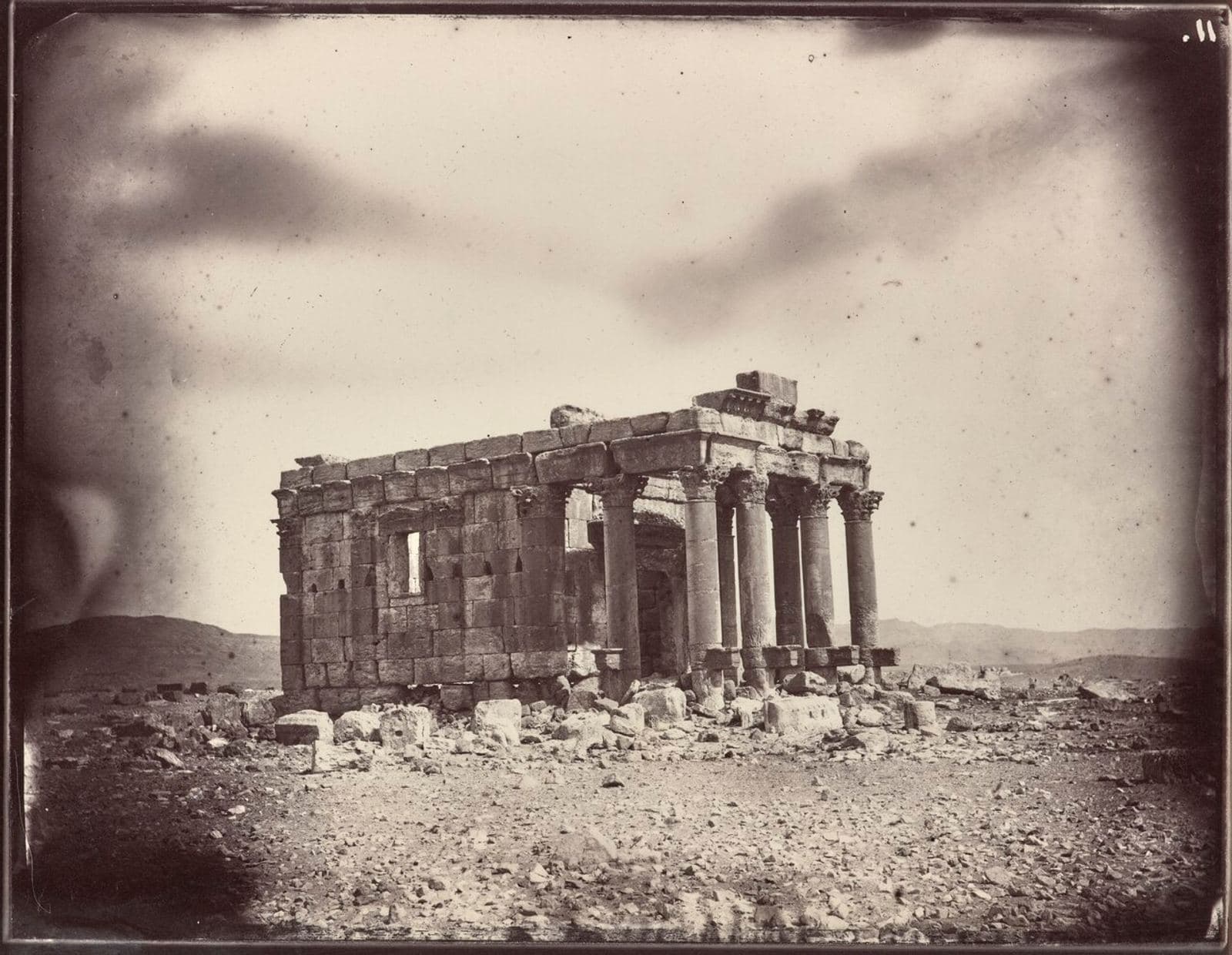 Sepia-toned 19th-century photograph of the ruined stone columns and stepped entrance of an ancient temple standing alone in a desert landscape