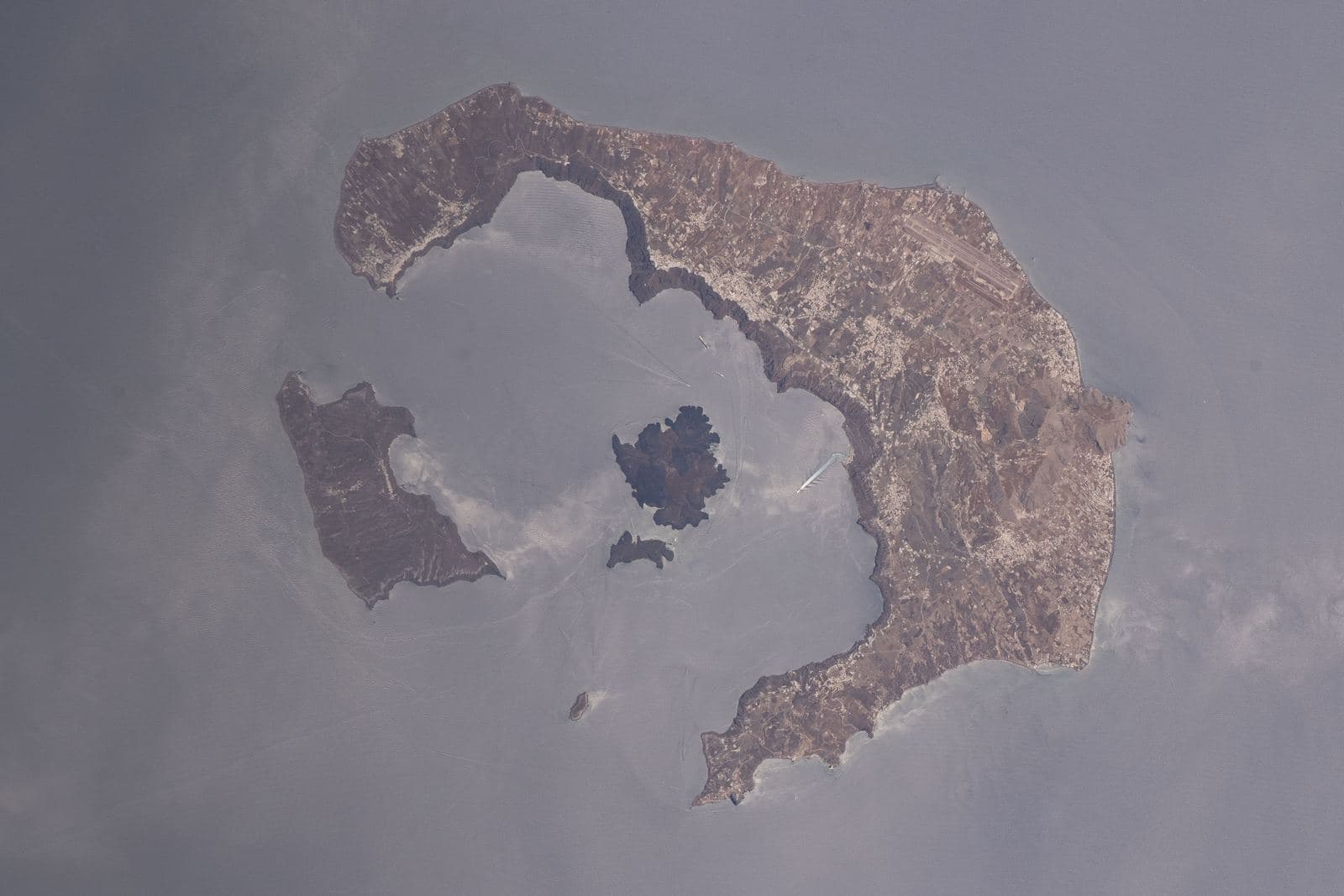 Photograph from orbit of a crescent-shaped island enclosing a flooded caldera with a dark volcanic islet at its centre